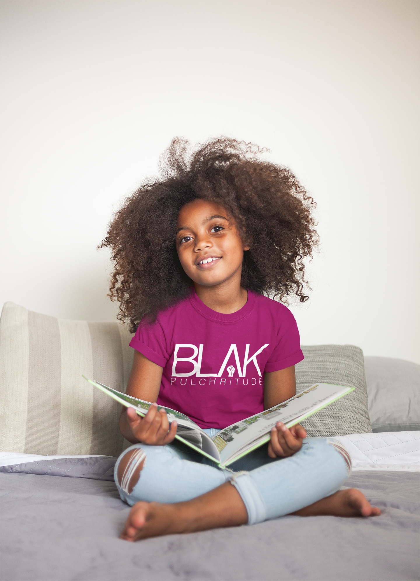 A young Black girl reading a book in her BLVK Pulchritude tee—playful and poised, embracing knowledge, joy, and style from the start.